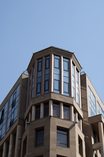 A modern building with a prominent octagonal section featuring vertical windows and columns. The façade is constructed with a combination of stone and glass, and the clear blue sky provides a stark background contrast.