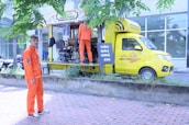 A mobile barbershop set up within a bright yellow truck is parked beside a sidewalk with two young men in vibrant orange uniforms. One man stands outside the truck on the pavement, and the other is inside the truck, which has its side open displaying a barber chair and accessories. There are trees and a building with large windows in the background.