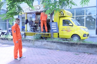 A mobile barbershop set up within a bright yellow truck is parked beside a sidewalk with two young men in vibrant orange uniforms. One man stands outside the truck on the pavement, and the other is inside the truck, which has its side open displaying a barber chair and accessories. There are trees and a building with large windows in the background.