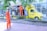 A mobile barbershop set up within a bright yellow truck is parked beside a sidewalk with two young men in vibrant orange uniforms. One man stands outside the truck on the pavement, and the other is inside the truck, which has its side open displaying a barber chair and accessories. There are trees and a building with large windows in the background.