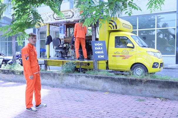 A mobile barbershop set up within a bright yellow truck is parked beside a sidewalk with two young men in vibrant orange uniforms. One man stands outside the truck on the pavement, and the other is inside the truck, which has its side open displaying a barber chair and accessories. There are trees and a building with large windows in the background.