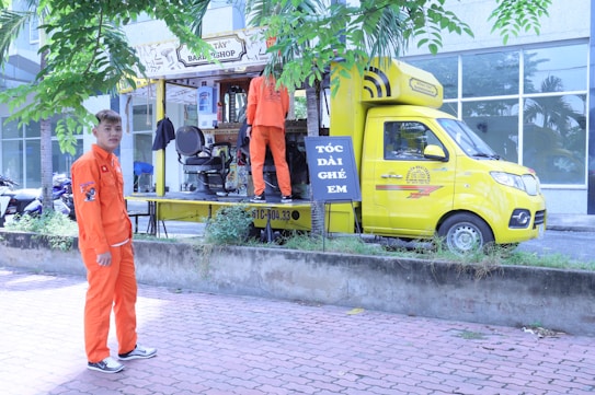 A mobile barbershop set up within a bright yellow truck is parked beside a sidewalk with two young men in vibrant orange uniforms. One man stands outside the truck on the pavement, and the other is inside the truck, which has its side open displaying a barber chair and accessories. There are trees and a building with large windows in the background.