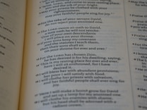 A close-up view of a page from a book displaying religious text. The text is formatted in verses, organized numerically, and printed in black ink on light-colored paper.