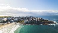 A coastal neighborhood is surrounded by the ocean on three sides. The cluster of buildings, primarily residential, sits on a rocky outcrop with waves gently lapping the shore. A sandy beach with visible people enjoying the water is prominent in the foreground. The sky features scattered clouds with patches of blue.