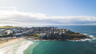 A coastal neighborhood is surrounded by the ocean on three sides. The cluster of buildings, primarily residential, sits on a rocky outcrop with waves gently lapping the shore. A sandy beach with visible people enjoying the water is prominent in the foreground. The sky features scattered clouds with patches of blue.