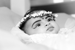A close-up black and white portrait of a baby wearing a floral headband, lying down. The baby has expressive eyes and a small bindi on the forehead.