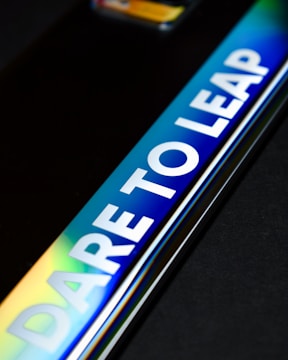 A close-up of a reflective metallic surface with the words 'DARE TO LEAP' prominently displayed in bold white letters. The surface shows a gradient of colors including blue, green, and yellow.