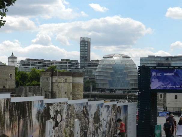 A skyline featuring modern and historic architecture including a modern glass building with a distinctive rounded shape. Historical stone towers and walls are visible in the foreground. A public path with people walking and reading signs is present. The cities' urban landscape is set under a partly cloudy sky.