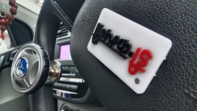 A close-up image of a car steering wheel adorned with a spinner knob on the left, which features a brand logo in blue. A white rectangular tag with some letters in black and red is attached to the steering wheel. The background shows part of the dashboard and a string of beads hanging from the rear-view mirror.