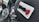 A close-up image of a car steering wheel adorned with a spinner knob on the left, which features a brand logo in blue. A white rectangular tag with some letters in black and red is attached to the steering wheel. The background shows part of the dashboard and a string of beads hanging from the rear-view mirror.