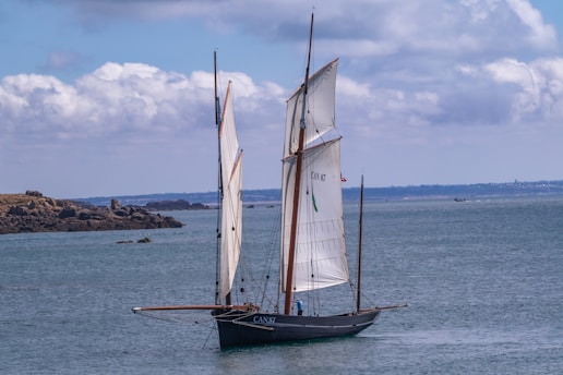 A large sailboat with two tall sails glides smoothly on calm blue waters. The boat is dark in color, possibly black, and displays the number CAN 87 on its sail. In the background, a rocky shoreline is visible under a partially cloudy sky.