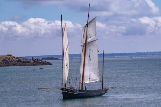 A large sailboat with two tall sails glides smoothly on calm blue waters. The boat is dark in color, possibly black, and displays the number CAN 87 on its sail. In the background, a rocky shoreline is visible under a partially cloudy sky.