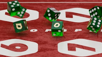 Green translucent dice with white dots appear to be in mid-air above a red surface with large white numbers and letters. The dice show different numbers on each face and have a glossy texture.