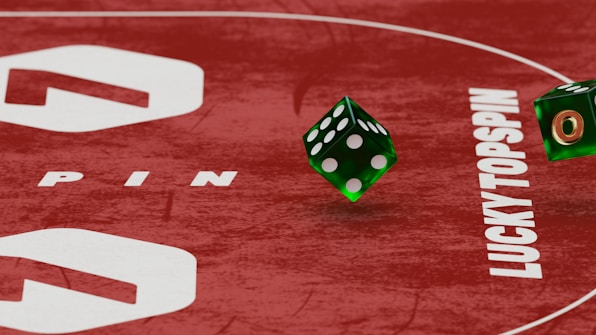 Two green dice with rounded edges are positioned on a red surface that seems to be part of a game board. The surface has white text and numbers on it, including the word 'LUCKYTOPSPIN' in capital letters.