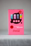 A promotional cardboard display features four collectible Coca-Cola glasses with various colors and designs. The background is bright pink, with bold red accents emphasizing 'APP EXCLUSIVE' and 'EXCLUSIVE TO McDonald’s'. The iconic Coca-Cola logo is prominently displayed.
