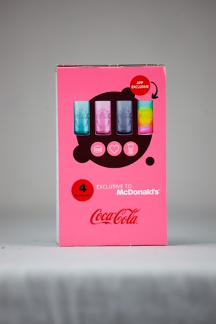 A promotional cardboard display features four collectible Coca-Cola glasses with various colors and designs. The background is bright pink, with bold red accents emphasizing 'APP EXCLUSIVE' and 'EXCLUSIVE TO McDonald’s'. The iconic Coca-Cola logo is prominently displayed.