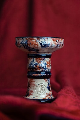 An intricately designed ceramic pedestal with a cylindrical shape. The surface features a textured pattern with splashes of blue and orange on a cream base. A subtle emblem is embossed near the bottom of the pedestal. A deep red fabric backdrop adds a rich contrast to the colors of the object.