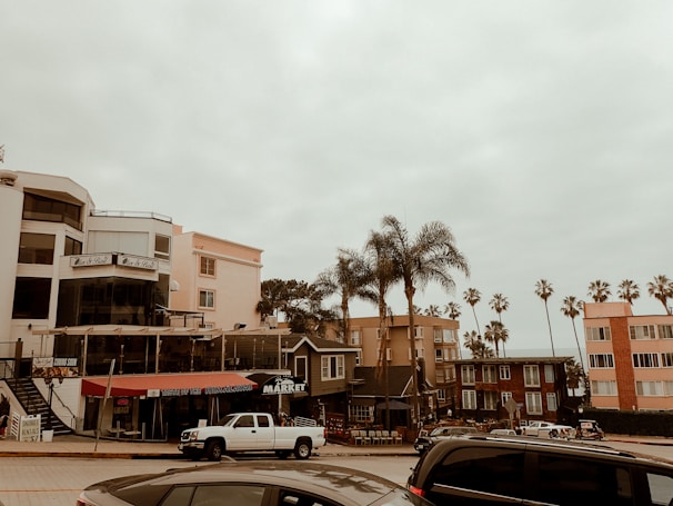 A coastal street scene featuring a row of buildings, including restaurants and a market. Tall palm trees are scattered among the buildings, and vehicles are parked along the street. The sky is overcast, giving the scene a muted ambiance.