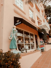 A street-level view of a shop with a large display window featuring various handbags and decorative items. A turquoise statue of a woman stands by the window, adorned with seashells and flowing garments. The building has a pink facade and a black awning with the label 'Norma Kay.' Potted plants and small shrubs line the walkway in front of the shop.