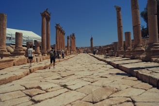 Ancient stone pillars line a wide, paved street, evoking a sense of historical grandeur. Several people are walking along this archaeological site, exploring the remnants of the past. The sky is clear and deep blue, contrasting the warm, earthy tones of the old stones.