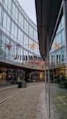 A modern architectural setting with glass walls reflecting the surroundings. Colorful butterfly decorations hang across the space above a cobblestone pathway, adding vibrant touches. Storefronts with visible signage and plants arranged in large pots decorate the walkway, creating a welcoming commercial atmosphere.