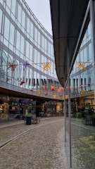 A modern architectural setting with glass walls reflecting the surroundings. Colorful butterfly decorations hang across the space above a cobblestone pathway, adding vibrant touches. Storefronts with visible signage and plants arranged in large pots decorate the walkway, creating a welcoming commercial atmosphere.