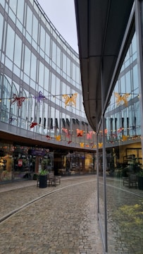 A modern architectural setting with glass walls reflecting the surroundings. Colorful butterfly decorations hang across the space above a cobblestone pathway, adding vibrant touches. Storefronts with visible signage and plants arranged in large pots decorate the walkway, creating a welcoming commercial atmosphere.