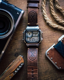 A digital watch with a leather strap is placed on a wooden surface. The watch displays the time and has a rugged design. Nearby, there is a vintage-style camera partially visible on the left, a corkscrew knife with a wooden handle below, and a braided rope on the right alongside a brown wallet.