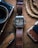 A digital watch with a leather strap is placed on a wooden surface. The watch displays the time and has a rugged design. Nearby, there is a vintage-style camera partially visible on the left, a corkscrew knife with a wooden handle below, and a braided rope on the right alongside a brown wallet.
