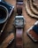 A digital watch with a leather strap is placed on a wooden surface. The watch displays the time and has a rugged design. Nearby, there is a vintage-style camera partially visible on the left, a corkscrew knife with a wooden handle below, and a braided rope on the right alongside a brown wallet.