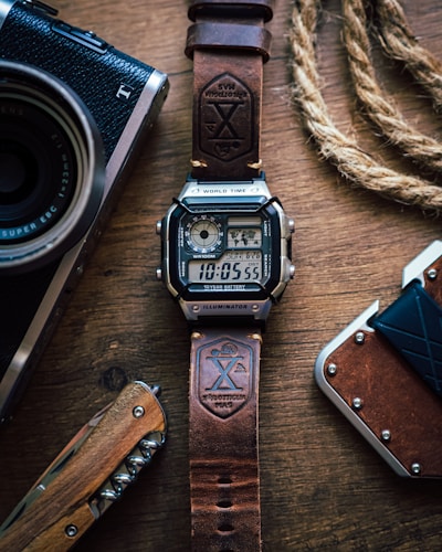 A digital watch with a leather strap is placed on a wooden surface. The watch displays the time and has a rugged design. Nearby, there is a vintage-style camera partially visible on the left, a corkscrew knife with a wooden handle below, and a braided rope on the right alongside a brown wallet.