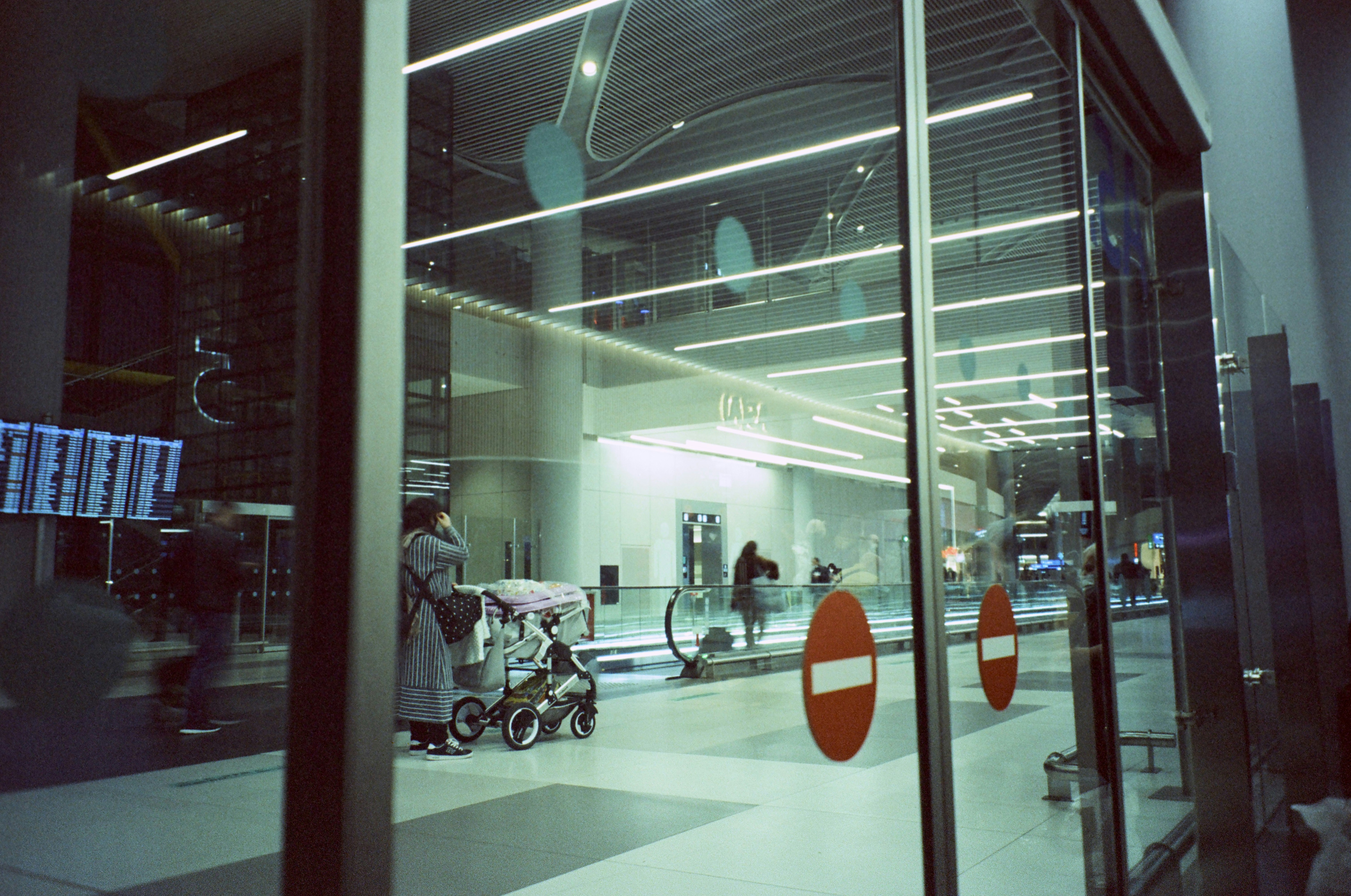 A modern airport terminal interior, viewed through glass doors marked with red 'no entry' signs. Inside, a person stands next to a stroller while travellers move along a travellator. The space is brightly lit with linear ceiling lights, and a digital flight information display is visible on the left.