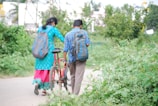 A man and a woman, both carrying backpacks, walk along a pathway surrounded by lush greenery. The woman is wearing a bright blue and pink traditional outfit, while the man is dressed in a blue plaid shirt and brown pants. They are accompanied by a red bicycle, which the man is guiding. The environment is serene with trees and plants on either side.