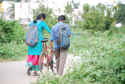 A man and a woman, both carrying backpacks, walk along a pathway surrounded by lush greenery. The woman is wearing a bright blue and pink traditional outfit, while the man is dressed in a blue plaid shirt and brown pants. They are accompanied by a red bicycle, which the man is guiding. The environment is serene with trees and plants on either side.