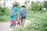 A man and a woman, both carrying backpacks, walk along a pathway surrounded by lush greenery. The woman is wearing a bright blue and pink traditional outfit, while the man is dressed in a blue plaid shirt and brown pants. They are accompanied by a red bicycle, which the man is guiding. The environment is serene with trees and plants on either side.
