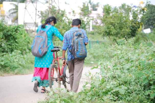 A man and a woman, both carrying backpacks, walk along a pathway surrounded by lush greenery. The woman is wearing a bright blue and pink traditional outfit, while the man is dressed in a blue plaid shirt and brown pants. They are accompanied by a red bicycle, which the man is guiding. The environment is serene with trees and plants on either side.