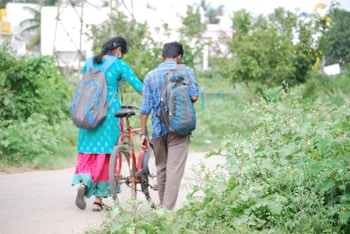 A man and a woman, both carrying backpacks, walk along a pathway surrounded by lush greenery. The woman is wearing a bright blue and pink traditional outfit, while the man is dressed in a blue plaid shirt and brown pants. They are accompanied by a red bicycle, which the man is guiding. The environment is serene with trees and plants on either side.