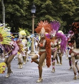 A vibrant parade featuring dancers in colorful feathered costumes, performing energetically on a cobblestone street. The background is lined with trees and a vintage street lamp.