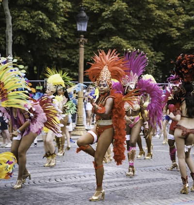 A vibrant parade featuring dancers in colorful feathered costumes, performing energetically on a cobblestone street. The background is lined with trees and a vintage street lamp.