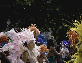 A vibrant outdoor carnival or parade scene featuring people dressed in elaborate, colorful costumes with feathers and headdresses. The participants are engaging in festivities, with a backdrop of trees and a crowd.