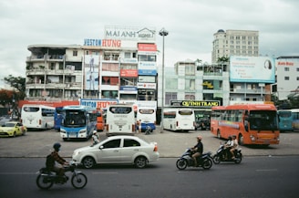 An urban scene featuring a busy street with cars and motorcycles in motion. In the background, there are multiple buildings with colorful advertisements and signs. Several buses are parked in a lot in front of these buildings, and people are seen walking and riding motorcycles. The sky is overcast.