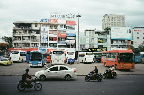 An urban scene featuring a busy street with cars and motorcycles in motion. In the background, there are multiple buildings with colorful advertisements and signs. Several buses are parked in a lot in front of these buildings, and people are seen walking and riding motorcycles. The sky is overcast.