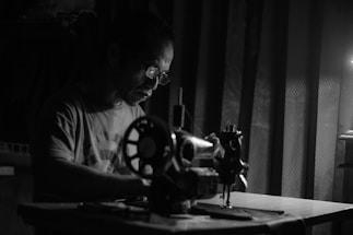 A man is concentrating intently as he works on a sewing machine. He is wearing glasses and a t-shirt, illuminated by a single light source, casting shadows around him. The setting is dimly lit, creating a dramatic contrast between light and dark areas.