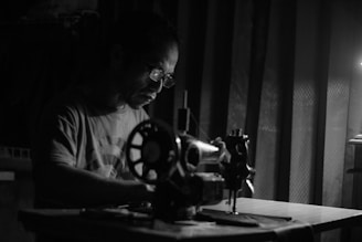 A man is concentrating intently as he works on a sewing machine. He is wearing glasses and a t-shirt, illuminated by a single light source, casting shadows around him. The setting is dimly lit, creating a dramatic contrast between light and dark areas.