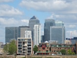 A cityscape featuring several modern skyscrapers with glass facades, including buildings labeled with names like JP Morgan, Citi, and HSBC. In the foreground, there are smaller brick buildings with traditional architecture, trees, and a clear blue sky in the background with scattered clouds.