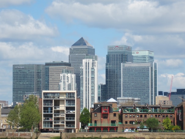 A cityscape featuring several modern skyscrapers with glass facades, including buildings labeled with names like JP Morgan, Citi, and HSBC. In the foreground, there are smaller brick buildings with traditional architecture, trees, and a clear blue sky in the background with scattered clouds.