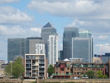 A cityscape featuring several modern skyscrapers with glass facades, including buildings labeled with names like JP Morgan, Citi, and HSBC. In the foreground, there are smaller brick buildings with traditional architecture, trees, and a clear blue sky in the background with scattered clouds.