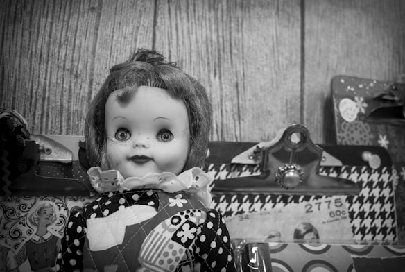 A vintage doll with curly hair and large eyes wearing a patterned dress, surrounded by clipboards and patterned materials, set against a wooden background.