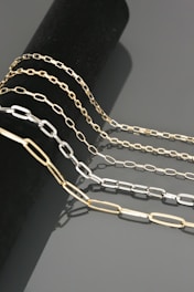 Four chains of varying lengths and colors drape elegantly over a cylindrical black surface. The chains have a sleek, metallic finish and are composed of rectangular links. The reflections on the smooth gray background add to the luxurious appearance.