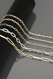 Four chains of varying lengths and colors drape elegantly over a cylindrical black surface. The chains have a sleek, metallic finish and are composed of rectangular links. The reflections on the smooth gray background add to the luxurious appearance.